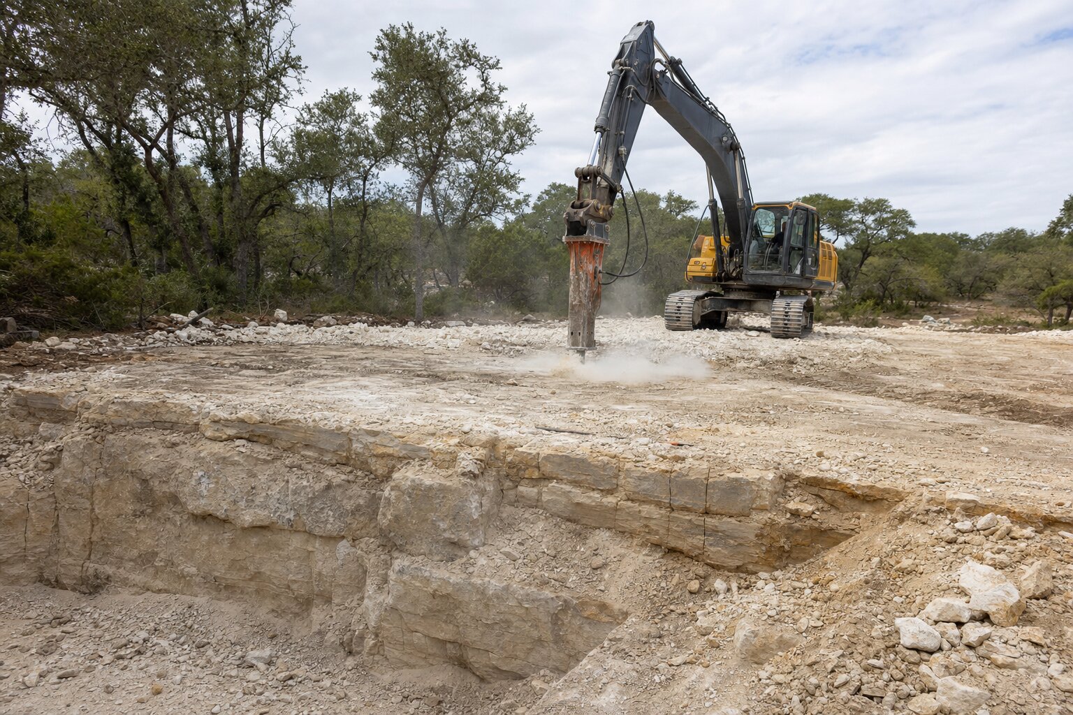 Excavator with a hydraulic rock breaker cutting a building pad into layered Texas Hill Country limestone bedrock