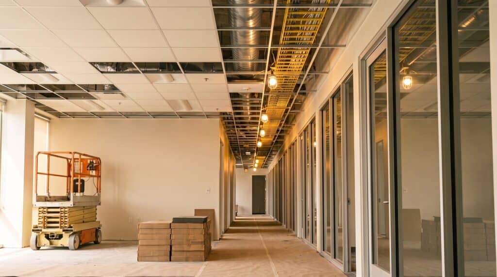 Office build-out in progress with a partly installed ceiling grid, overhead cable tray and ductwork, and glass-front private offices